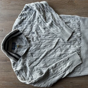 Grå stickad tröja från Polo Ralph Lauren - Säljer en snygg grå stickad tröja från Polo Ralph Lauren. Den har ett klassiskt kabelstickat mönster och en ribbad krage med svarta och grå ränder. Perfekt för höst och vinter, med långa ärmar och ribbade muddar för extra komfort. Passar både till vardags och lite finare tillfällen.
