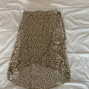 Leopardmönstrad kjol från KappAhl - Säljer en superfin leopardmönstrad kjol från KappAhl. Kjolen har en omlott-design med knytband i midjan, vilket ger en snygg och justerbar passform. Den är i en beige färg med svarta leopardfläckar, perfekt för både vardag och fest. Materialet känns lätt och luftigt, vilket gör den idealisk för varmare dagar.