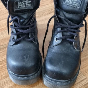 Svarta kängor med stålhätta från Dr. Martens - Säljer ett par svarta kängor från Dr. Martens i skinn med stålhätta. De har en robust sula och snörning framtill. Perfekta för höst och vinter, dessa kängor är både stiliga och praktiska.