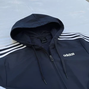 Adidas vindjacka med luva blå - HELT NY. Adidas, tunnare mörkblå sportig vindjacka som passar bra för utomhusaktiviteter när det är vindigt. Kan användas som uppvärmingsplagg innan en fotbollsträning eller liknande.                             Butikpris: 1 199kr