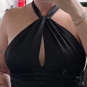 Svart halterneck topp - Snygg svart halterneck topp med en twist-detalj framtill. Toppen är ärmlös och har en öppen rygg, perfekt för festliga tillfällen. Materialet ser ut att vara mjukt och stretchigt för en bekväm passform.