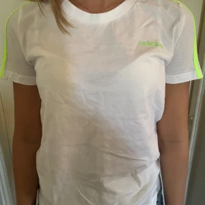 Vit t-shirt från Adidas - Säljer en snygg vit t-shirt från Adidas med neongröna detaljer. Den har korta ärmar och en slim passform, perfekt för en sportig look. T-shirten är i nyskick och har fortfarande prislappen kvar. 
