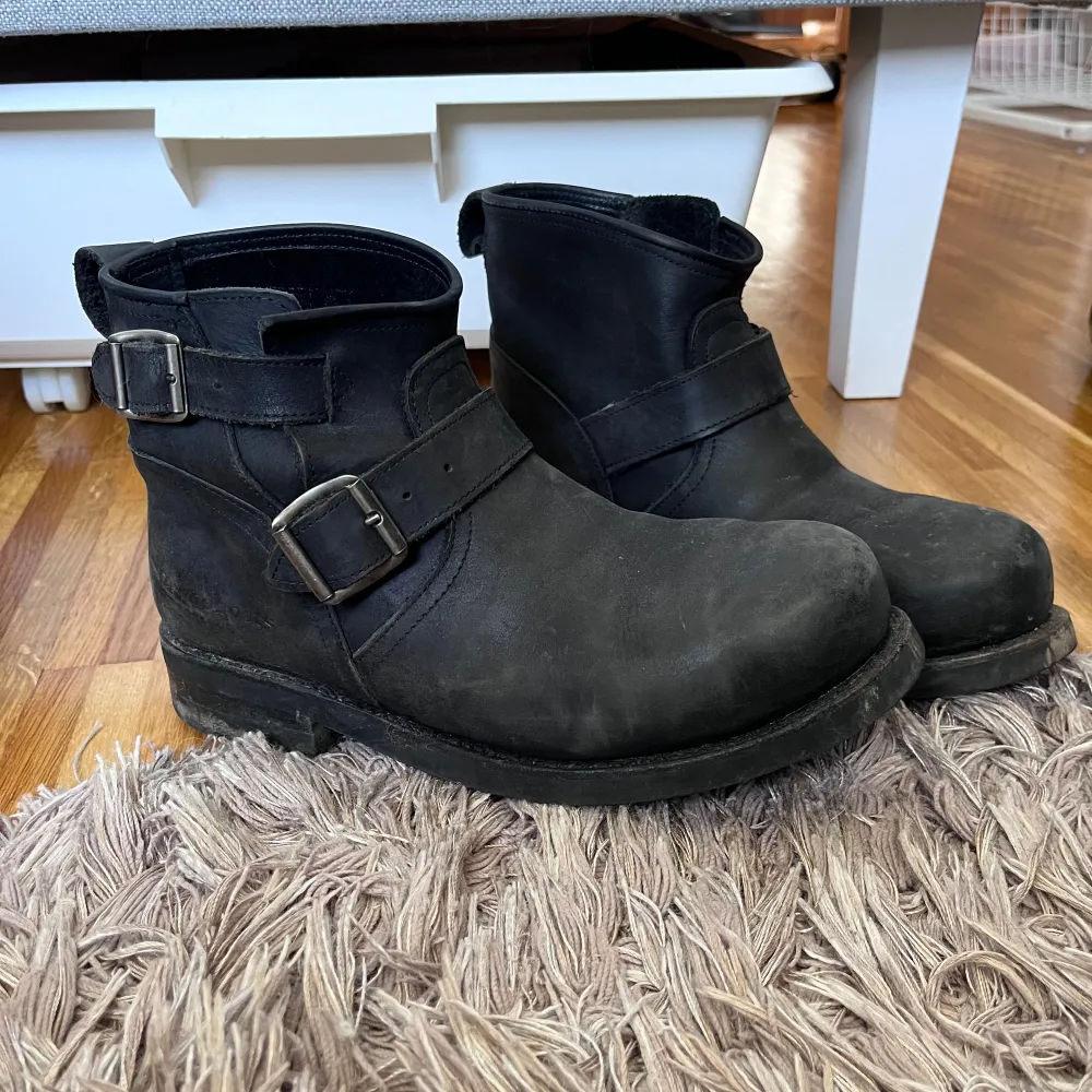 Säljer ett par svarta boots i skinn med coola spännen på sidan. De har en robust sula som är oljebeständig, perfekt för höst och vinter. Skorna är välanvända men fortfarande i bra skick. Perfekta för en tuff och stilren look!. Skor.