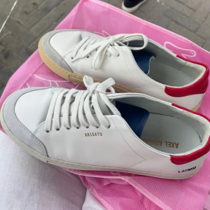 Nya Vita sneakers från Axel Arigato - Säljer ett par snygga vita sneakers från Axel Arigato. De har en stilren design med röd detalj vid hälen och en beige sula. Skorna är i skinn och har snörning framtill. Perfekta för både vardag och fest!