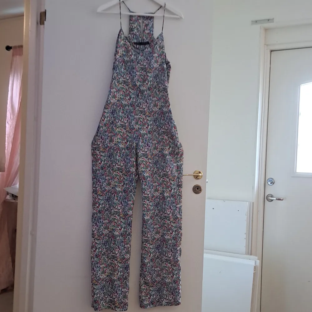 Säljer en superfin blommig jumpsuit med smala axelband. Perfekt för sommardagar eller en avslappnad fest. Det har en lös passform och är gjord i ett lätt och bekvämt material. Den har ett färgglatt mönster som verkligen sticker ut!. Jeans & Byxor.