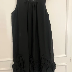 Svart ärmlös klänning med volangdetaljer -  svart ärmlös klänning med vackra volangdetaljer längs nederkanten. Luftig passform och tillverkad i ett lätt material. Unik och finns ej att köpa liknande