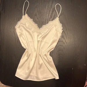 Vit satinlinne med spetsdetaljer från bikbok  - Elegant vit satinlinne med smala axelband och vacker spets vid halsringningen. Perfekt för en stilren och sofistikerad look.
