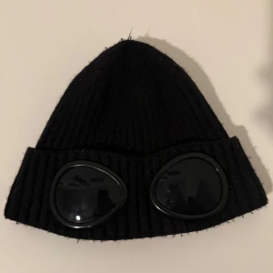 Cp Company Mössa - Snygg svart ribbad mössa från C.P. Company med unika glasögondetaljer framtill. Perfekt för att ge din outfit en cool touch. Tillverkad i ett bekvämt material som håller dig varm.