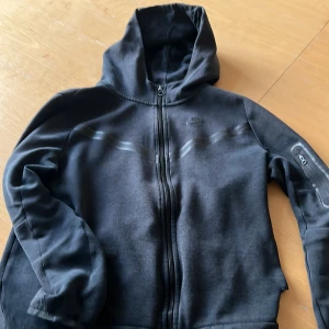 Svart hoodie från Nike - Säljer en svart hoodie från Nike med dragkedja och huva. Perfekt för en avslappnad stil. Den har en ficka på ärmen och är tillverkad i ett mjukt material. Priset kan sänkas också!