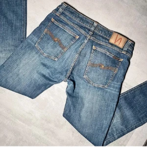Blå jeans från Nudie Jeans - Snygga blå jeans från Nudie Jeans med klassisk femficksdesign och orange sömmar. De har en knappgylf och är tillverkade i slitstarkt denim. Perfekta för en avslappnad stil. Storlek 32 nyrpris runt 2000