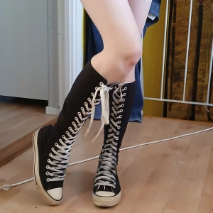 Knähöga convers Svarta höga sneakers med snörning - Snygga svarta höga sneakers med vit snörning som går hela vägen upp till knäna. Perfekta för en cool och avslappnad stil. De har en klassisk vit sula och tåhätta som ger en retro känsla.