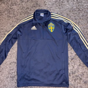Sverige blå  träningsjacka från Adidas   - Säljer en blå träningsjacka från Adidas med Sveriges emblem på bröstet. Jackan har en halv dragkedja och de klassiska Adidas-ränderna i vitt längs ärmarna. Perfekt för fotbollsträning eller som en sportig vardagsjacka.