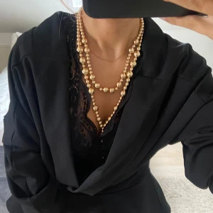 Guldigt halsband med pärlor - Elegant guldigt halsband med dubbla rader av glänsande pärlor. Perfekt för att ge en lyxig touch till din outfit. Halsbandet har en vacker droppformad detalj som hänger nedtill.
