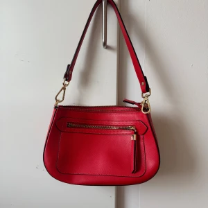 Röd axelväska med dragkedja - Snygg röd axelväska i syntetmaterial med guldfärgade detaljer. Väskan har en praktisk dragkedja framtill och en justerbar axelrem. Perfekt för att ge en färgklick till din outfit. Nypris 399