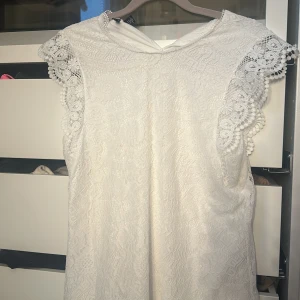 Vit spets topp - Elegant vit blus med vacker spetsdetaljering. Toppen  är ärmlös och har en fin struktur i tyget som ger en romantisk känsla. Perfekt för speciella tillfällen.