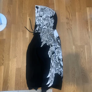 Svart hoodie med dödskalle och tigertryck - Cool svart hoodie med ett stort vitt tryck av en dödskalle och en tiger. Hoodien har en justerbar huva med snören.