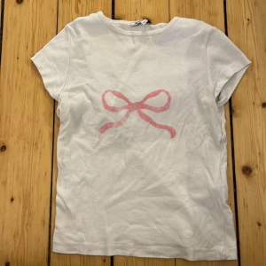 Vit t-shirt med rosa rosett från subdued - Säljer en söt vit t-shirt från subdued med en rosa rosett tryckt på framsidan. T-shirten har korta ärmar och är tillverkad i ett mjukt bomullsmaterial. Perfekt för en avslappnad stil. Orginalpris över 300kr.