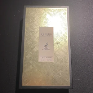 Domino Essence Eau de Parfum - Upptäck Domino Essence från Maison Alhambra, en elegant Eau de Parfum med en sofistikerad doft. Flaskan har en stilren design med en gyllene etikett och svart lock. Perfekt för den som söker en unik och minnesvärd doftupplevelse.