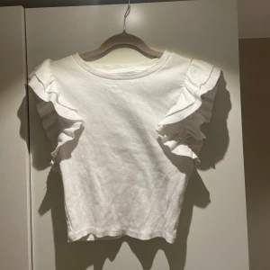 Vit ribbad topp från Zara - Säljer en söt vit ribbad topp från Zara med volangdetaljer på ärmarna. Perfekt för en stilren look. Toppen har en rund halsringning och är tillverkad i ett mjukt material.