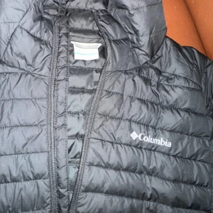 Svart pufferjacka från Columbia - Säljer en svart pufferjacka från Columbia med dragkedja och huva. Jackan är quiltad och perfekt för kyligare väder. Den har en klassisk design med Columbia-loggan på bröstet.