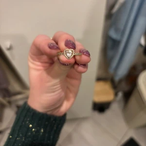 Pandora Guldfärgad ring med hjärtformad sten - Elegant guldfärgad ring med en stor hjärtformad sten i mitten, omgiven av mindre stenar. Perfekt för att ge en romantisk touch till din stil.