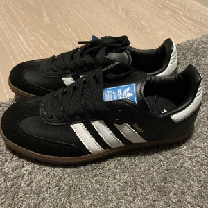 Adidas Samba OG i svart och vit - Säljer ett par klassiska Adidas Samba OG sneakers i svart och vit. Skorna har en stilren design med de ikoniska tre ränderna och en brun sula. Perfekta för en sportig och trendig look.