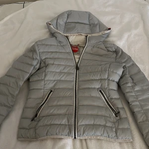 Grå dunjacka från Colmar - Snygg och stilren grå dunjacka från Colmar med huva och dragkedja. Jackan har två praktiska fickor med dragkedjor och en logotyp på ärmen. Perfekt för hösten och våren!💓