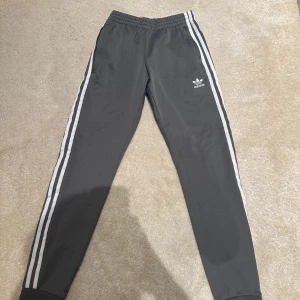 Grå byxor från Adidas - Snygga grå byxor från Adidas med de klassiska tre vita ränderna längs sidorna. Byxorna har en elastisk midja för extra komfort. Koftan finns till dessa byxor så skriv i dm om ni vill ha båda samtidigt så löser jag ett bättre pris.