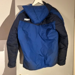Blå och svart pufferjacka från Puma - Säljer en blå och svart pufferjacka från Puma med huva och dragkedja. Jackan har långa ärmar och är perfekt för kyligare väder. Den har en sportig look med Puma-loggan på bröstet.