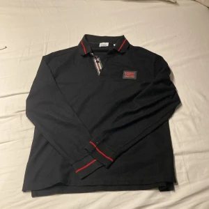 Svart pikétröja från Burberry - Snygg svart pikétröja från Burberry med röda detaljer på kragen och ärmsluten. Tröjan har en klassisk krage och knappar framtill med ett mönstrat inslag. Burberry-logga på bröstet ger en exklusiv touch.