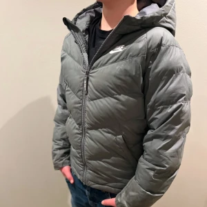 Grå dunjacka från Nike - Snygg grå dunjacka från Nike med huva och dragkedja. Jackan har en pufferdesign och är perfekt för kyligare väder. Den har långa ärmar och en diskret logga på bröstet.