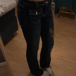 Mörkblå jeans med fickdetaljer - Snygga mörkblå jeans med unika fickdetaljer framtill. De har en låg midja och en utsvängd passform som ger en klassisk look. Perfekta för en avslappnad stil.