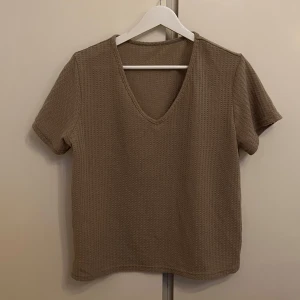 Brun ribbad t-shirt - Säljer en stilren ljusbrun t-shirt med v-ringning och korta ärmar. Den har en ribbad textur som ger en extra touch till designen. Perfekt för en avslappnad look.