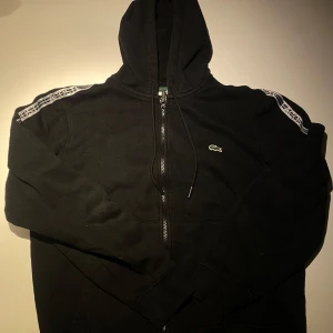 Svart hoodie från Lacoste - Snygg svart hoodie från Lacoste med dragkedja och huva. Den har vita detaljer längs ärmarna och den klassiska krokodilloggan på bröstet. Perfekt för en avslappnad stil. Passar både M och S
