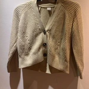 Beige stickad kofta från Lindsey Kids - Säljer en mysig beige stickad kofta från Lindsey Kids. Koftan har en v-ringad design med knappar framtill och ribbade detaljer. Perfekt för kyliga dagar!