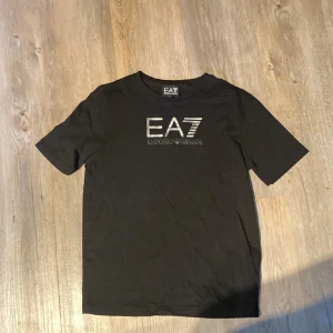 Svart t-shirt från Emporio Armani - Snygg svart t-shirt från Emporio Armani med EA7-logga på bröstet. Perfekt för en stilren look. Tillverkad i mjukt material för extra komfort. Passar utmärkt till både jeans och shorts.