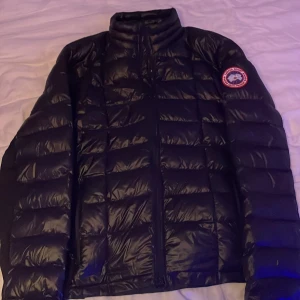 Svart dunjacka från Canada Goose - Säljer en svart dunjacka från Canada Goose med deras ikoniska logga på ärmen. Jackan har en quiltad design och dragkedja framtill. Perfekt för kyliga dagar. Och har även ett litet hål i armen som man inte ens ser. Där priset även blir ganska billigt på grund av att jag haft de n länge liggandes i garderoben. Storleken är xl men sitter perfekt för L
