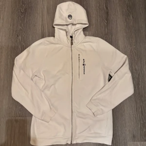Sail Racing zip hoodie  - Säljer en stilren vit huvtröja från Sail Racing med dragkedja framtill. Tröjan har en justerbar huva och långa ärmar. Perfekt för en avslappnad stil.