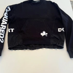 Svart tröja från DSQUARED2 - Säljer en svart tröja från DSQUARED2 med tryck på ärmarna och en stor ficka framtill. Tröjan har elastisk nederkant och texten 'DOMINATE YOUR SPORT'. Använd 2 ggr, nypris 5000:-