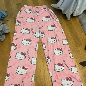 Rosa Hello Kitty pyjamasbyxor - Säljer ett par rosa pyjamasbyxor med Hello Kitty-motiv. Byxorna har ett elastiskt midjeband och är dekorerade med söta Hello Kitty-ansikten och text. Perfekta för en mysig hemmakväll!