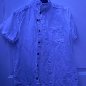 Vit kortärmad skjorta från KappAhl - Säljer en stilren vit kortärmad skjorta från KappAhl. Skjortan har en ståkrage och knappar framtill. Perfekt för en avslappnad och snygg look. 🕶️