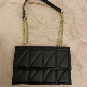 Svart quiltad axelväska med kedja - Snygg svart axelväska i quiltat mönster med guldfärgad kedja som axelrem. Väskan har en stilren design med magnetlås och gott om plats för dina nödvändigheter. Perfekt för att ge din outfit en elegant touch. Passar både till vardag och fest! 