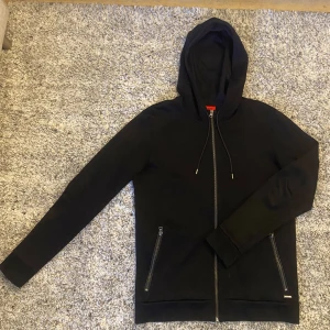 Svart hoodie från Hugo Boss - Säljer en stilren svart hoodie från Hugo Boss med dragkedja och justerbar huva. Tröjan har långa ärmar och praktiska fickor med dragkedja. Perfekt för en avslappnad look. Hoodie är i ett något tunnare tygmaterial som andas bra och därmed är perfekt som undertröja eller på en varm vår/höst-dag!  1999:- nypris. Om du önskar ytterligare info eller specifika bilder så är det bara att skriva 🙏