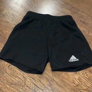 Svarta träningsshorts från Adidas - Säljer ett par svarta träningsshorts från Adidas. De har en elastisk midja och är perfekta för sport och träning. Adidas-loggan syns tydligt på framsidan. Passar bra för aktiva dagar!