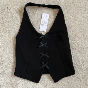 Svart väst med rosetter M oanvänd  - Snygg svart väst med dekorativa rosetter framtill. Perfekt för att ge en stilren touch till din outfit. Västen har en v-ringad design och är i en klassisk svart färg.