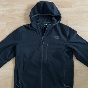 Svart North face vindjacka  - Säljer en stilren svart vindjacka från The North Face. Jackan har en praktisk huva och dragkedja framtill. Perfekt för blåsiga dagar och har en enkel design med The North Face-logga på bröstet.