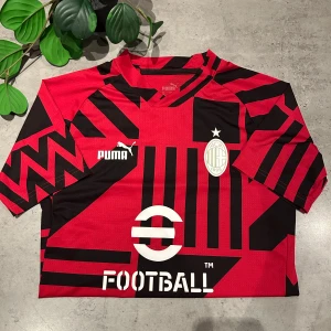 Röd och svart fotbollströja från Puma - Snygg röd och svart fotbollströja från Puma med ett iögonfallande geometriskt mönster. Tröjan har korta ärmar och ett klubbmärke på bröstet. Perfekt för fotbollsfans som vill sticka ut på planen.