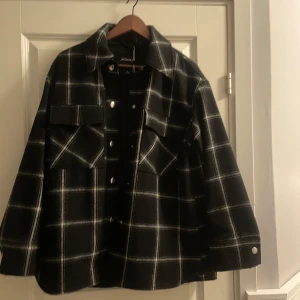 Svart och vit rutig jacka från Monki - Snygg svart och vit rutig jacka från Monki med knappar framtill och två bröstfickor. Jackan har en klassisk krage och långa ärmar. Perfekt för en stilren look.