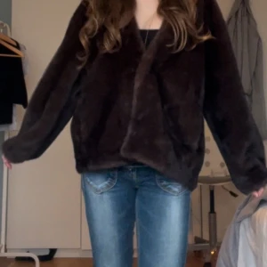 Brun pälsjacka - Säljer en mysig brun pälsjacka med långärmad design. Jackan har en enkel och stilren look utan synliga detaljer som knappar eller dragkedja. Perfekt för att hålla sig varm och snygg under kyliga dagar.