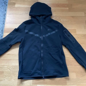 Svart hoodie från Nike - Säljer en svart hoodie från Nike med dragkedja och huva. Jackan har en diskret Nike-logga på bröstet och en praktisk ficka med dragkedja på ärmen. Perfekt för en sportig look.